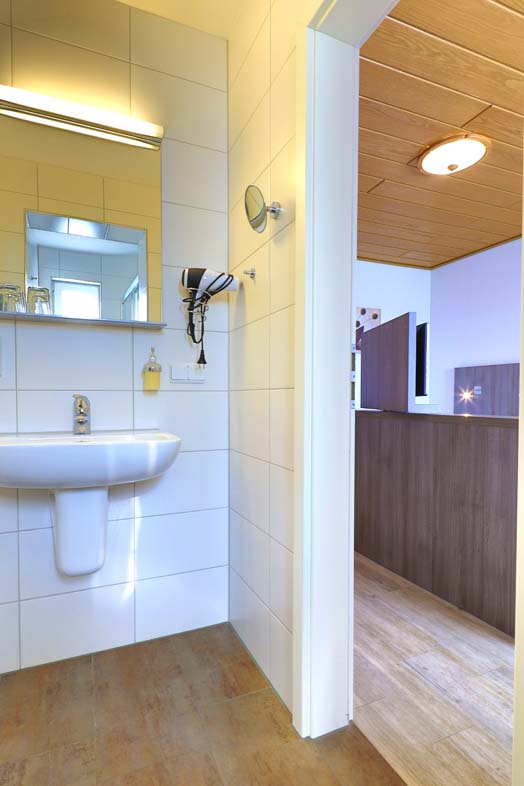 Badezimmer mit weißem Waschbecke, Föhn, Kosmetikspiegel und beleuchteter Wandspiegel, Seifenspender, die Tür steht offen