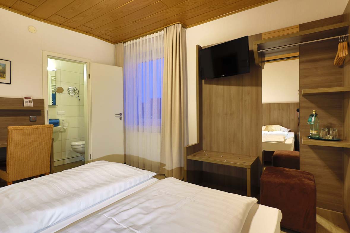Zimmer mit weiß bezogenen Betten ein offener Einbauschrank mit integrietem TV neben einem Fenster mit Vorhängen. Alles in Erdtönen gehalten