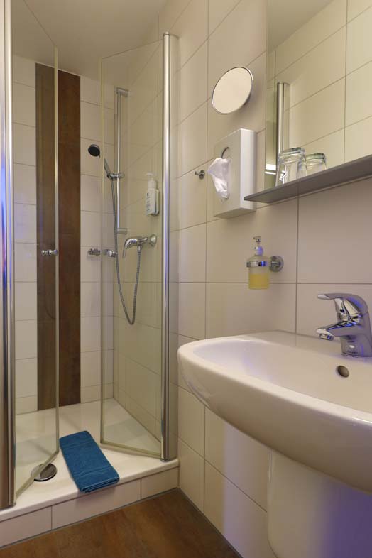 Ein Badezimmer mit Blick in die Dusche welche einen braunen laengsstreifen gefliesst hat am Wannenrand liegt eine petrolfarbene Duschvorlage