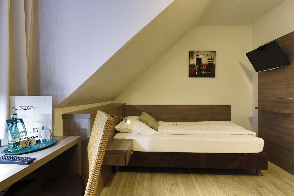 Ein Hotelzimmer mit Blick auf das Einzelbett, ein TV hängt am Fußende an der Wand, der Schreibtisch steht vor dem Fenster