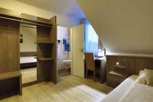 Ein Hotelzimmer mit Blick auf Schreibtisch und offener Schrankanlage, dieTür zum Badezimmer steht offen