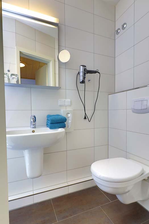 Bathroom with white wall tiles, brown floor tiles, petrol-colored towels lie on the washbasin, a hair dryer and cosmetic mirror hang next to the washbasin