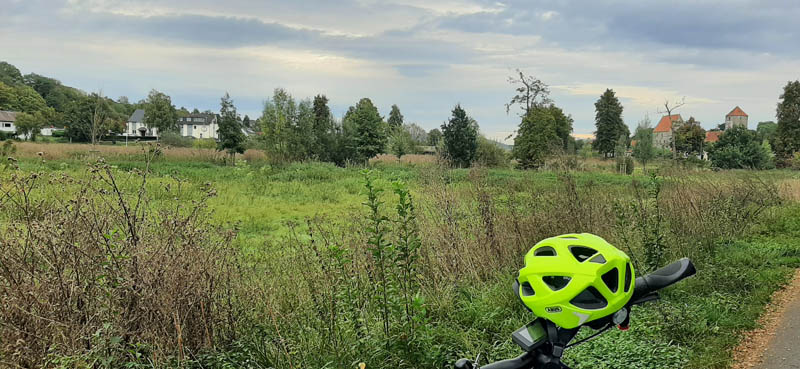 Wiese, im Hintergrund rechts den Landgasthof zur scharfen Ecke und links die Domäne Marienburg im Vorderen sieht man einen Neonegelben Fahrradhelm